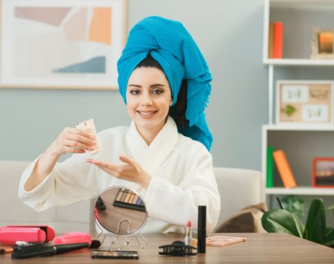 smiling-young-girl-holding-tone-up-cream-wrapped-hair-towel-sitting-table-with-makeup-tools-living-room (1)